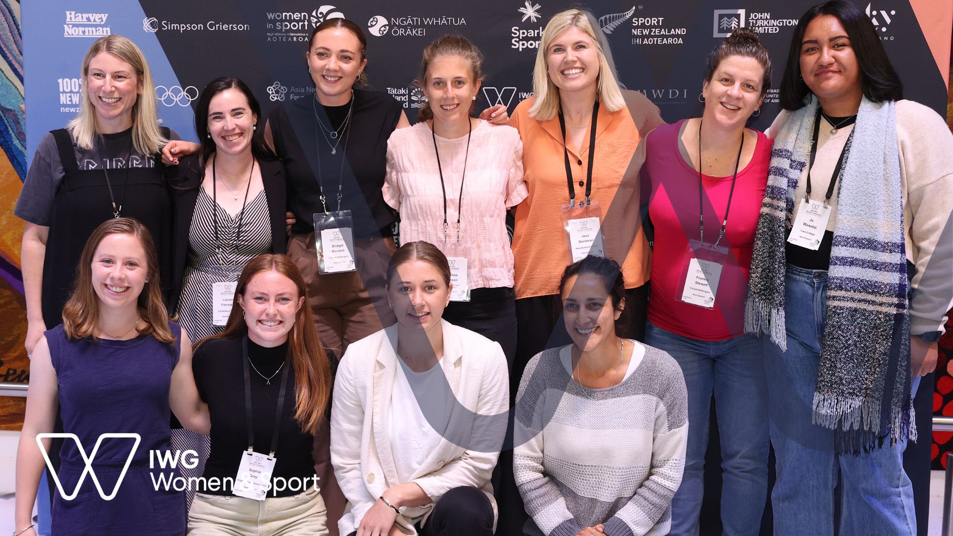 A large group of women stand against a branded backdrop and smile at the camera at the IWG Women and Sport Conference in New Zealand, 2022